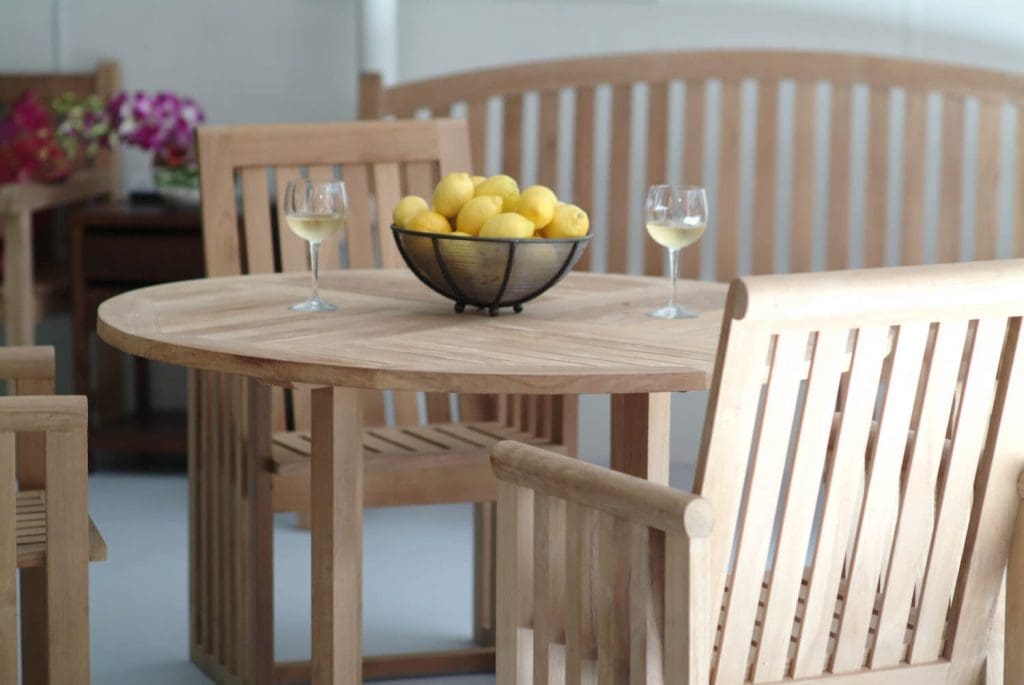 Teak patio furniture in backyard - a patio bench, 4 patio chairs, and a patio table - with 2 filled wine glasses and a basket of lemons on the table